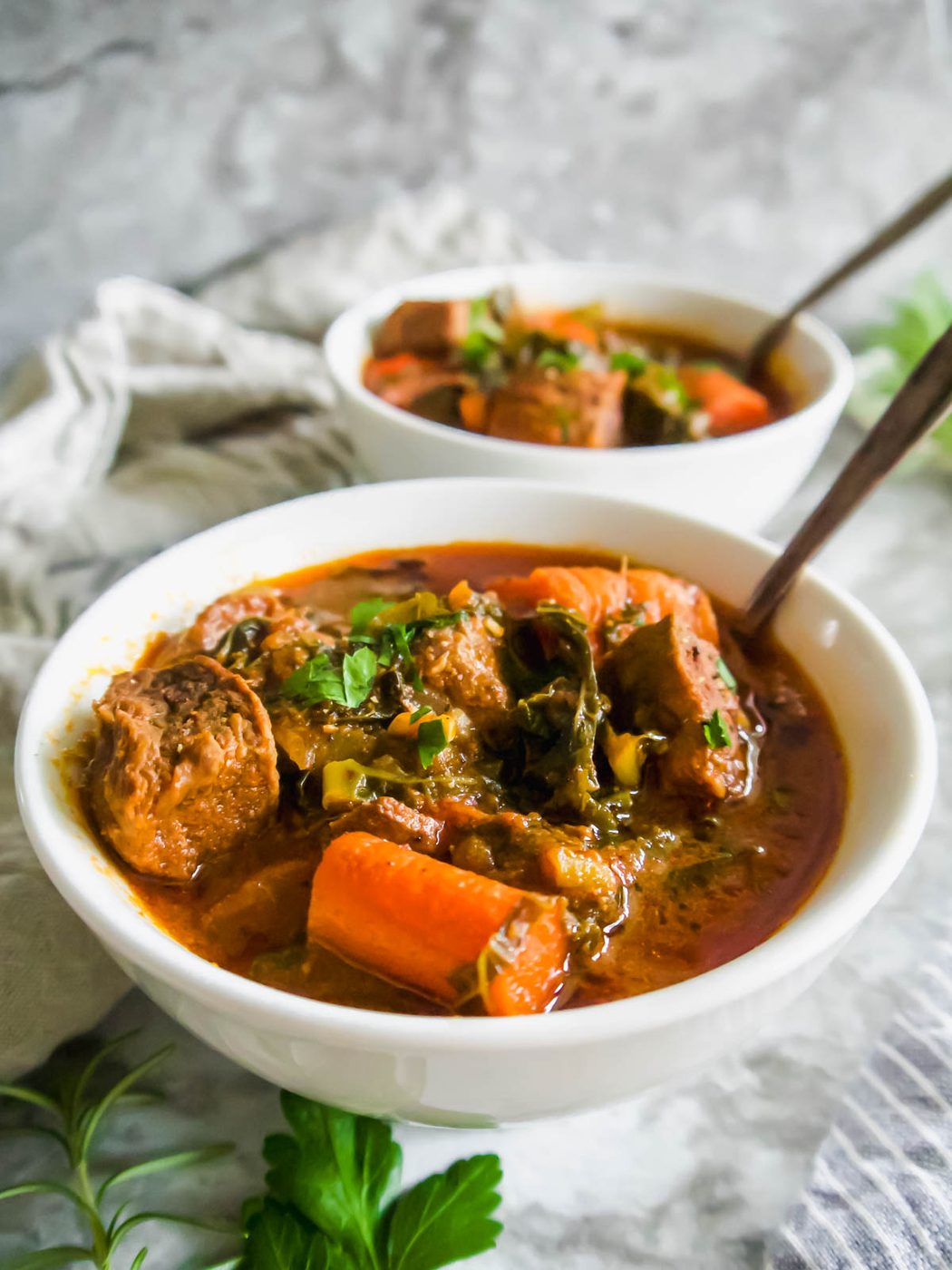 Side angle of beef and kale stew in two bowls with spoons inside them.