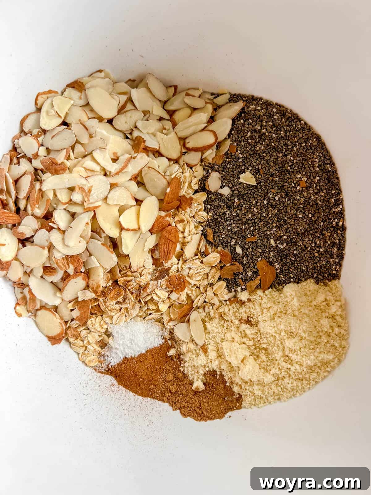 Artisan Olive Oil Granola Almonds and Chia 4 A large mixing bowl containing the dry ingredients for olive oil granola: rolled oats, almond flour, chia seeds, sliced almonds, cinnamon, nutmeg, and sea salt, awaiting mixing.