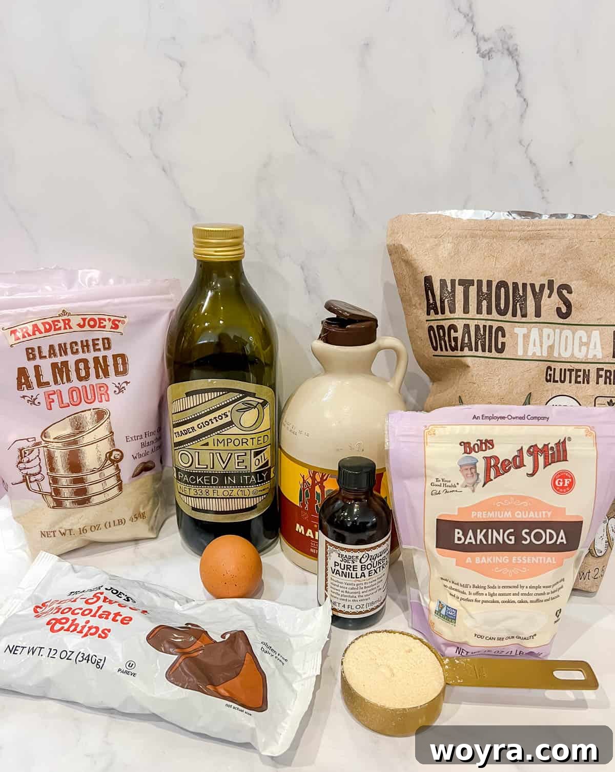 A flat lay of ingredients laid out on a surface, including olive oil, maple syrup, vanilla extract, an egg, various flours (almond, tapioca, coconut), baking soda, salt, and chocolate chips, ready for making paleo soft gluten-free chocolate chip cookies.