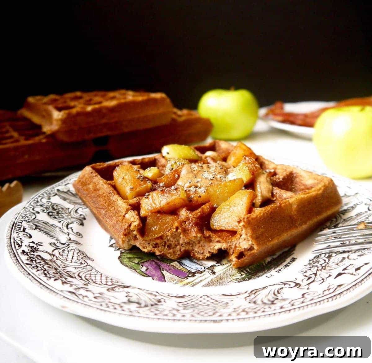 A close-up view of a gluten-free apple cinnamon waffle on a plate, showcasing its golden crispy exterior and a bite taken out, revealing the soft interior flecked with apple pieces.
