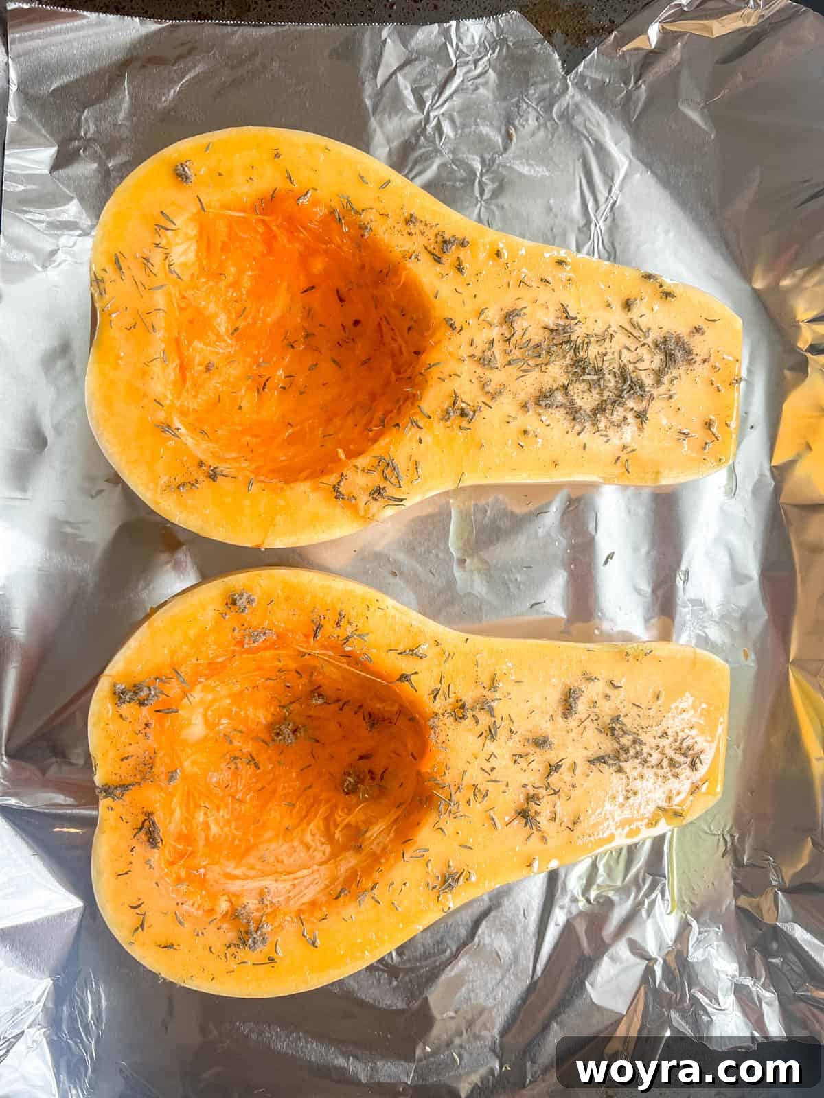 A butternut squash cut in half lengthwise with seeds removed, brushed with olive oil and herbs, ready for roasting.