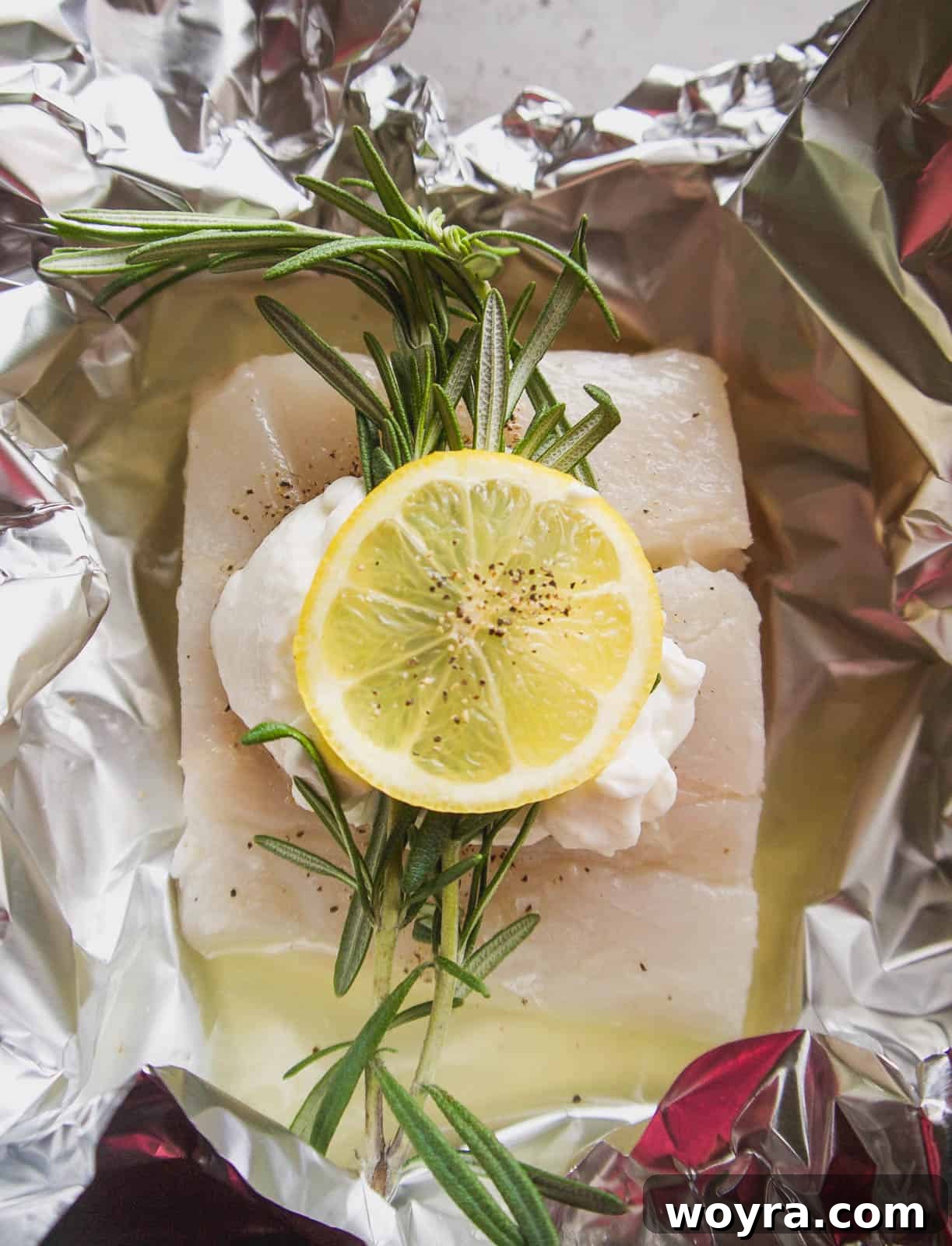 Halibut fillets seasoned with lemon, salt, and pepper, topped with Greek yogurt, rosemary, and lemon slices, ready to be wrapped in foil.