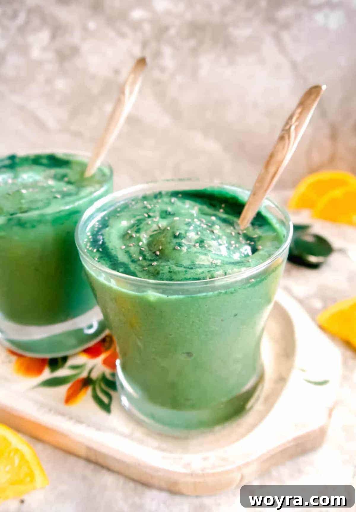 Spirulina Sunshine Smoothie 3 A side view of two cups filled with the vibrant green spirulina banana smoothie, with soft focus oranges positioned in the background, creating a fresh and inviting scene.