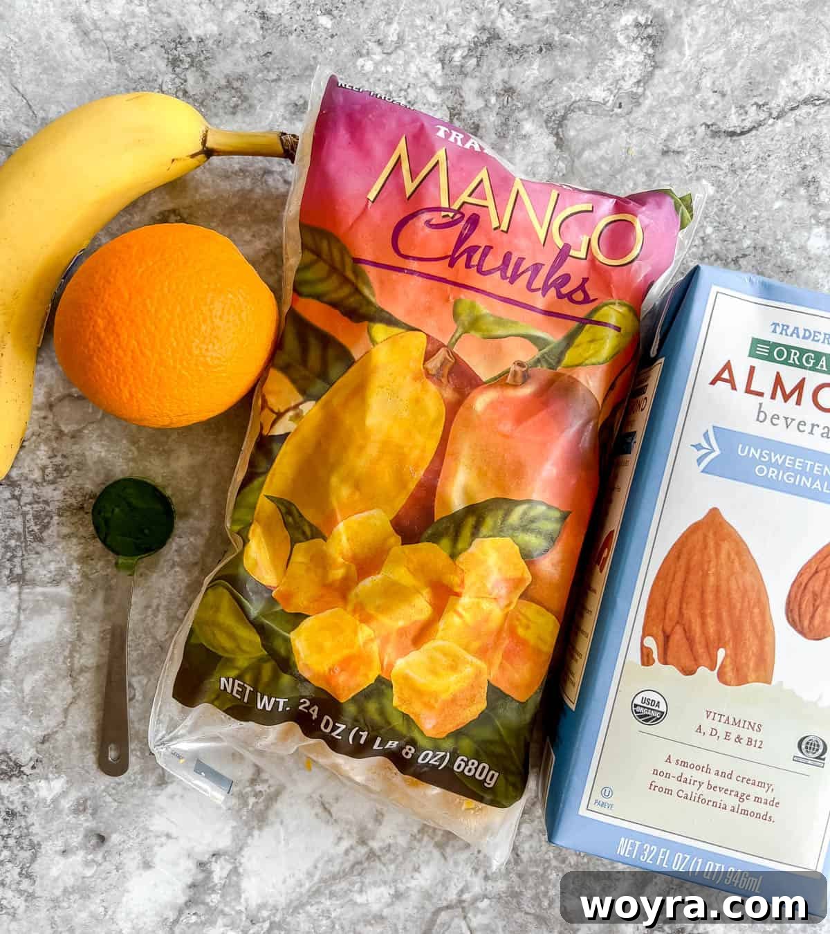 Spirulina Sunshine Smoothie 4 A neatly arranged selection of raw ingredients required for the spirulina smoothie, including a freshly peeled orange, generous chunks of frozen mango, a whole frozen banana, a vibrant green container of spirulina powder, and a carton of unsweetened almond milk.