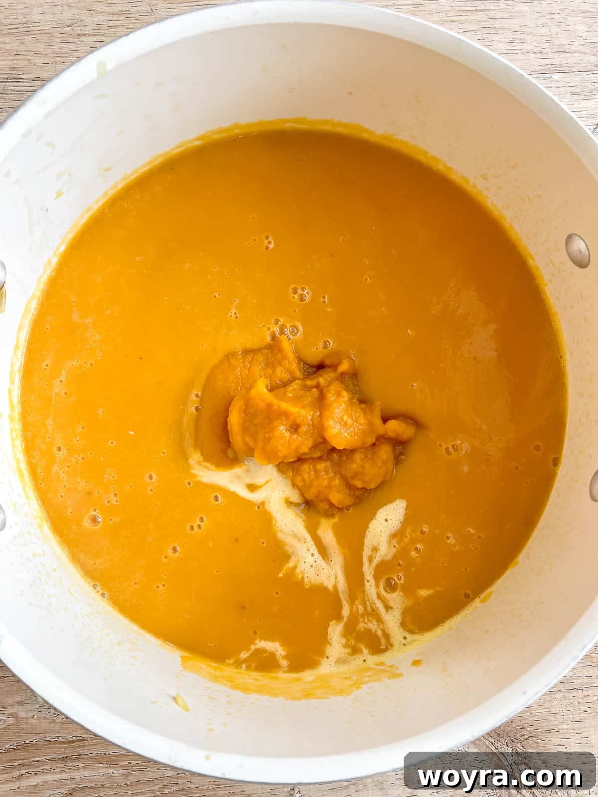 Pumpkin puree and heavy cream being stirred into the blended sweet potato soup in a pan, adding richness and color.