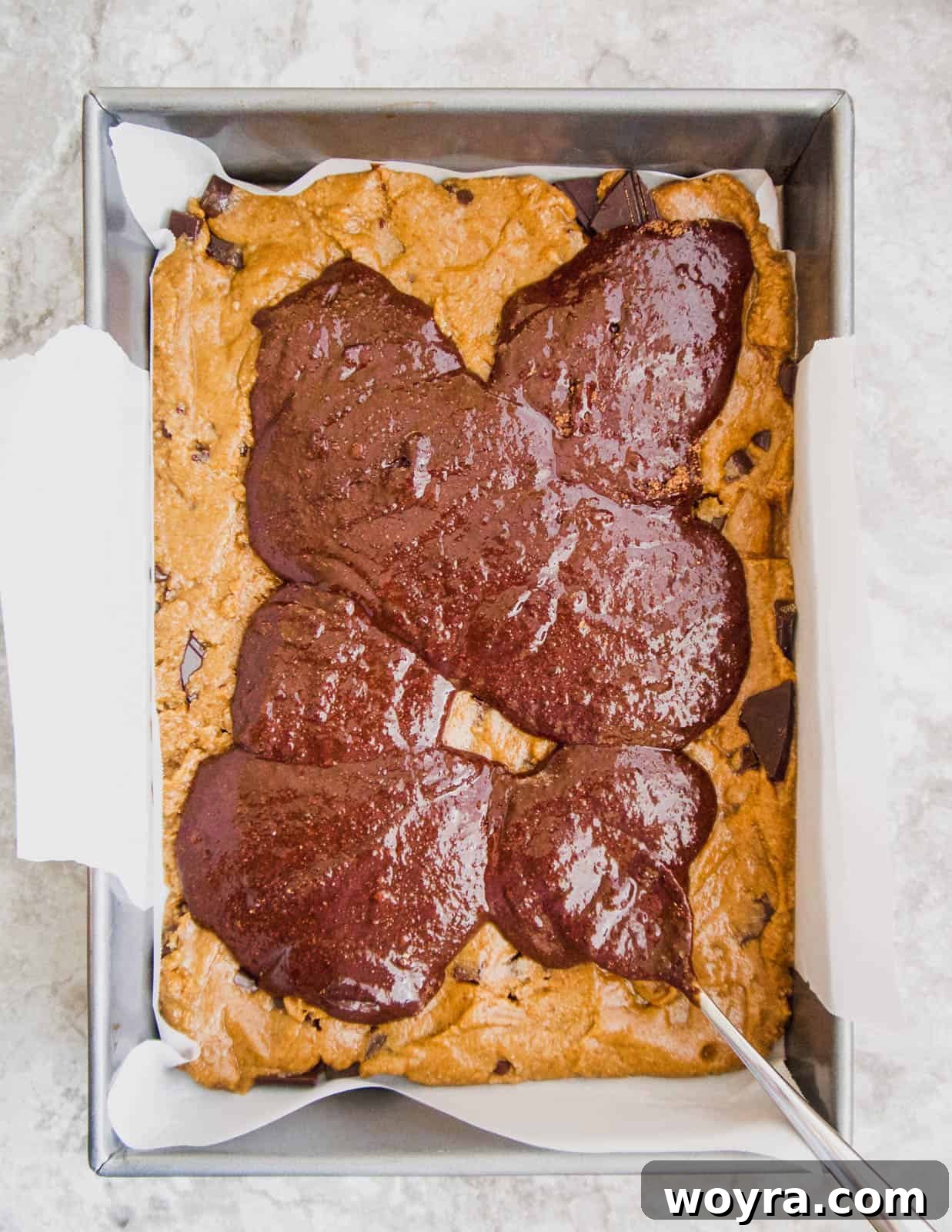 The brownie batter being carefully poured over the cookie layer in the baking pan.