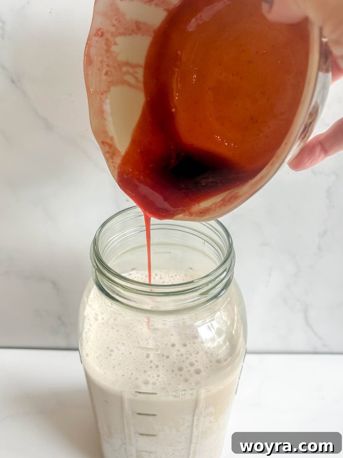 Homemade Strawberry Almond Milk: A Vegan Delight 13 The vibrant red strawberry syrup being carefully poured into the freshly made white almond milk in a glass container, before mixing.