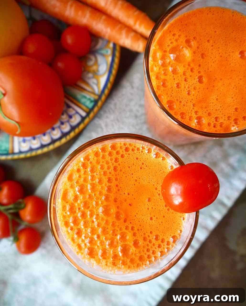 Rejuvenating Tomato Carrot Fusion 2 Aerial view of a tall glass filled with bright orange and red tomato carrot juice, garnished with a slice of orange and a carrot stick, sitting on a light surface.