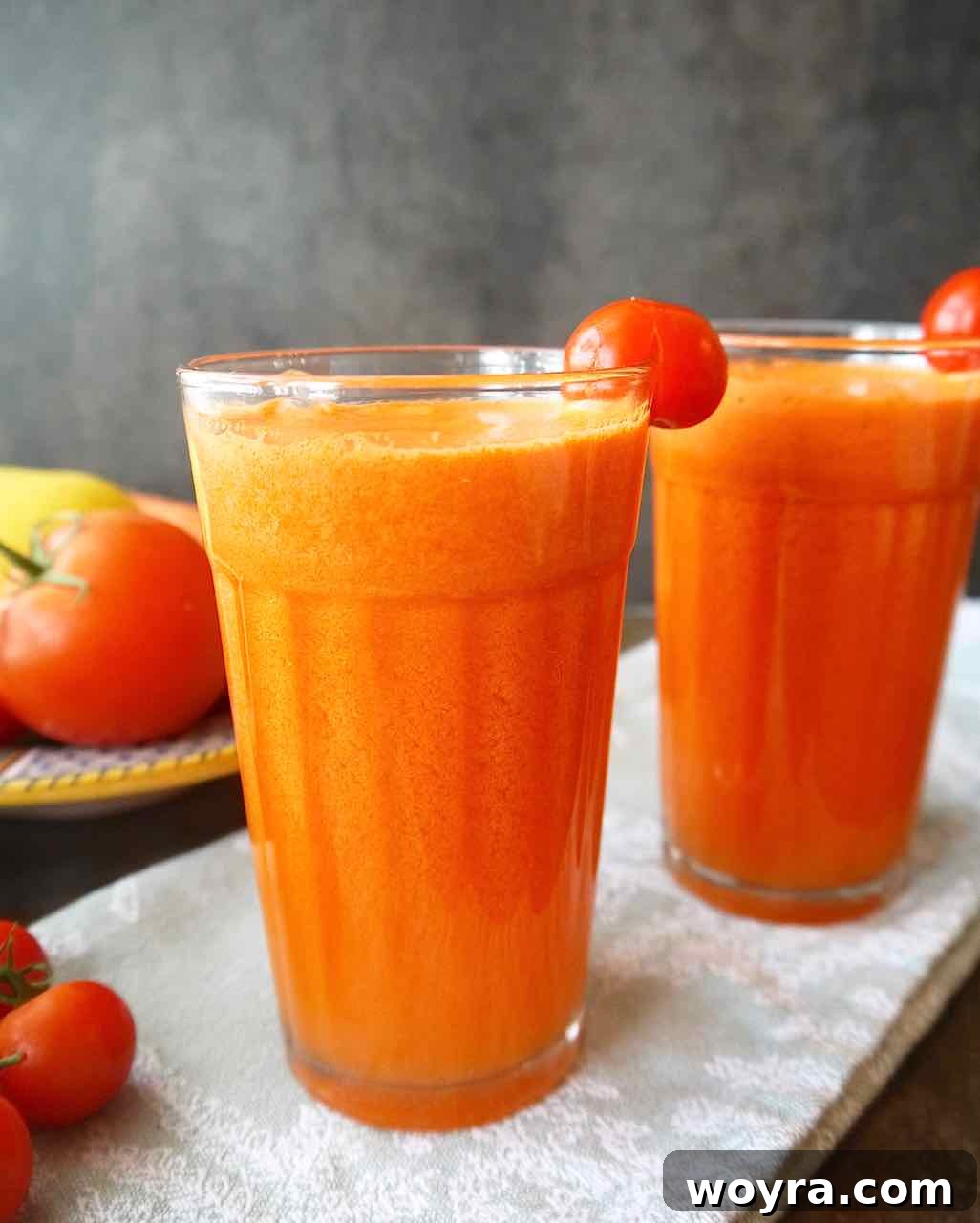 Rejuvenating Tomato Carrot Fusion 5 Two glasses of vibrant carrot tomato juice, one with a cherry tomato resting on the rim, served on a rustic wooden board.