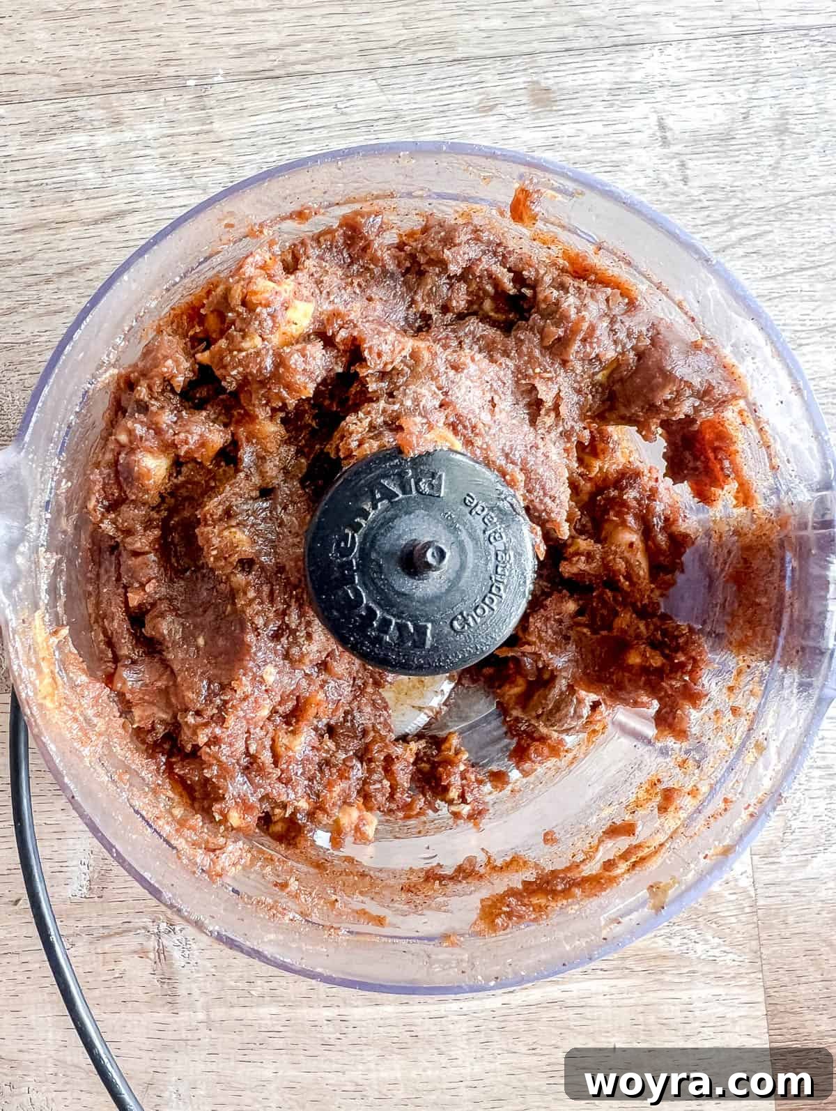 Date paste, maple syrup, vanilla extract, almond butter, cinnamon, nutmeg, and dried apples are being blended in a food processor.