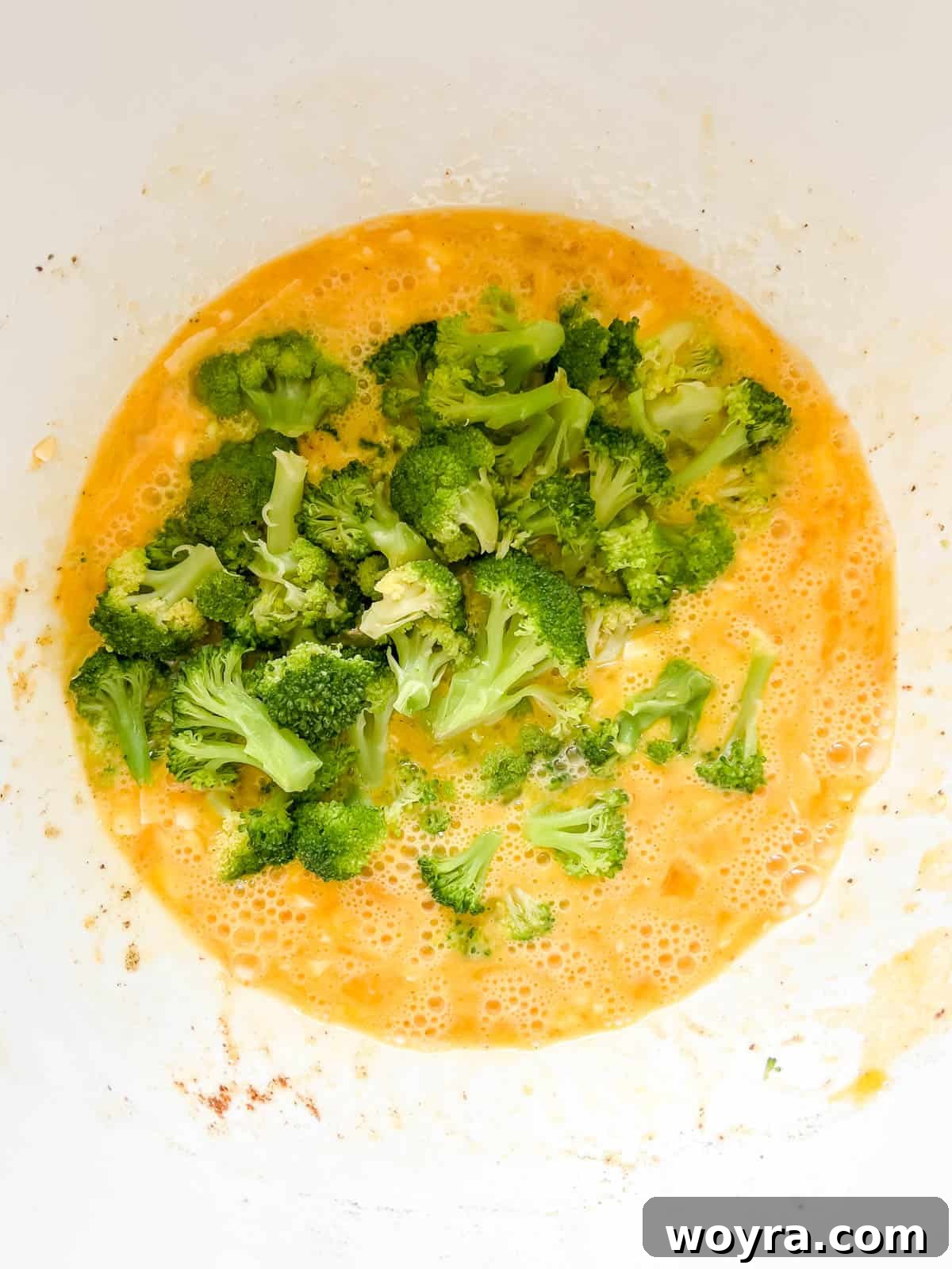 Steamed broccoli florets being folded into the egg and cheese mixture in a bowl.