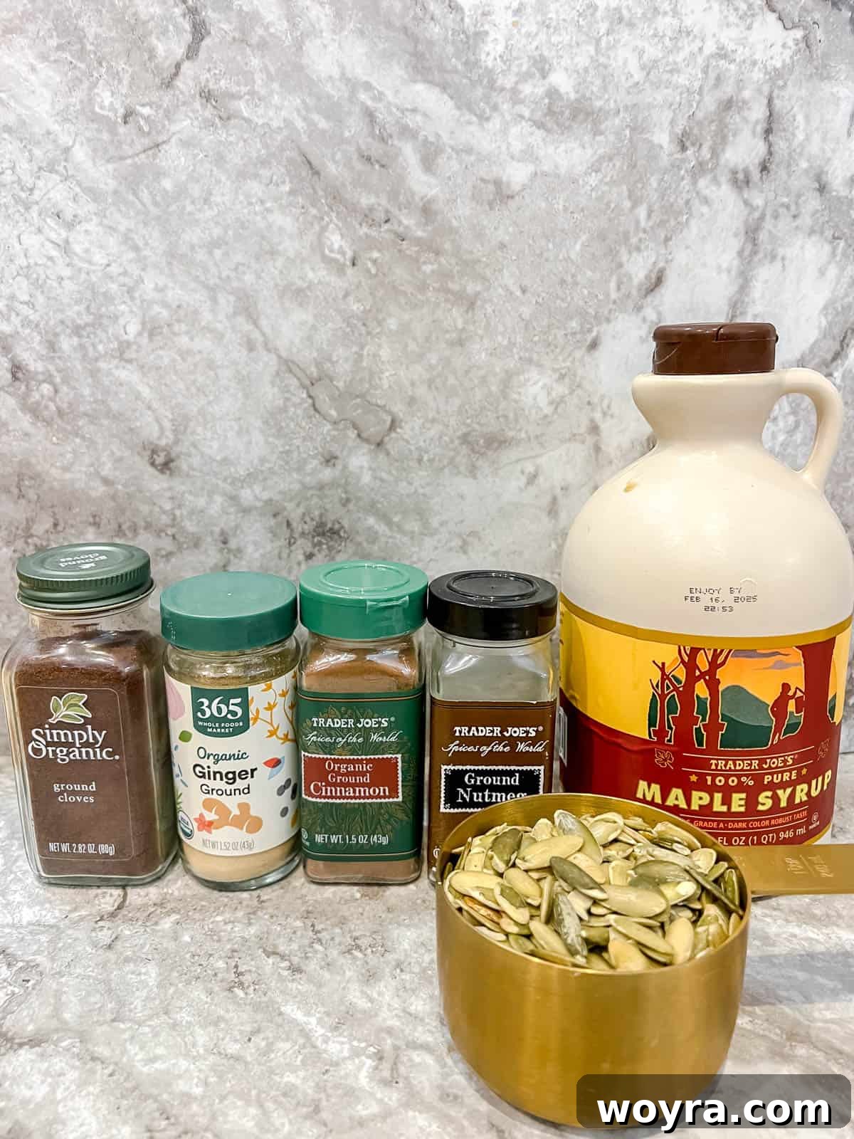 A flat lay photograph showcasing the ingredients for maple roasted pepitas: green pepitas, pure maple syrup, ground cinnamon, nutmeg, ginger, and cloves arranged on a neutral surface.
