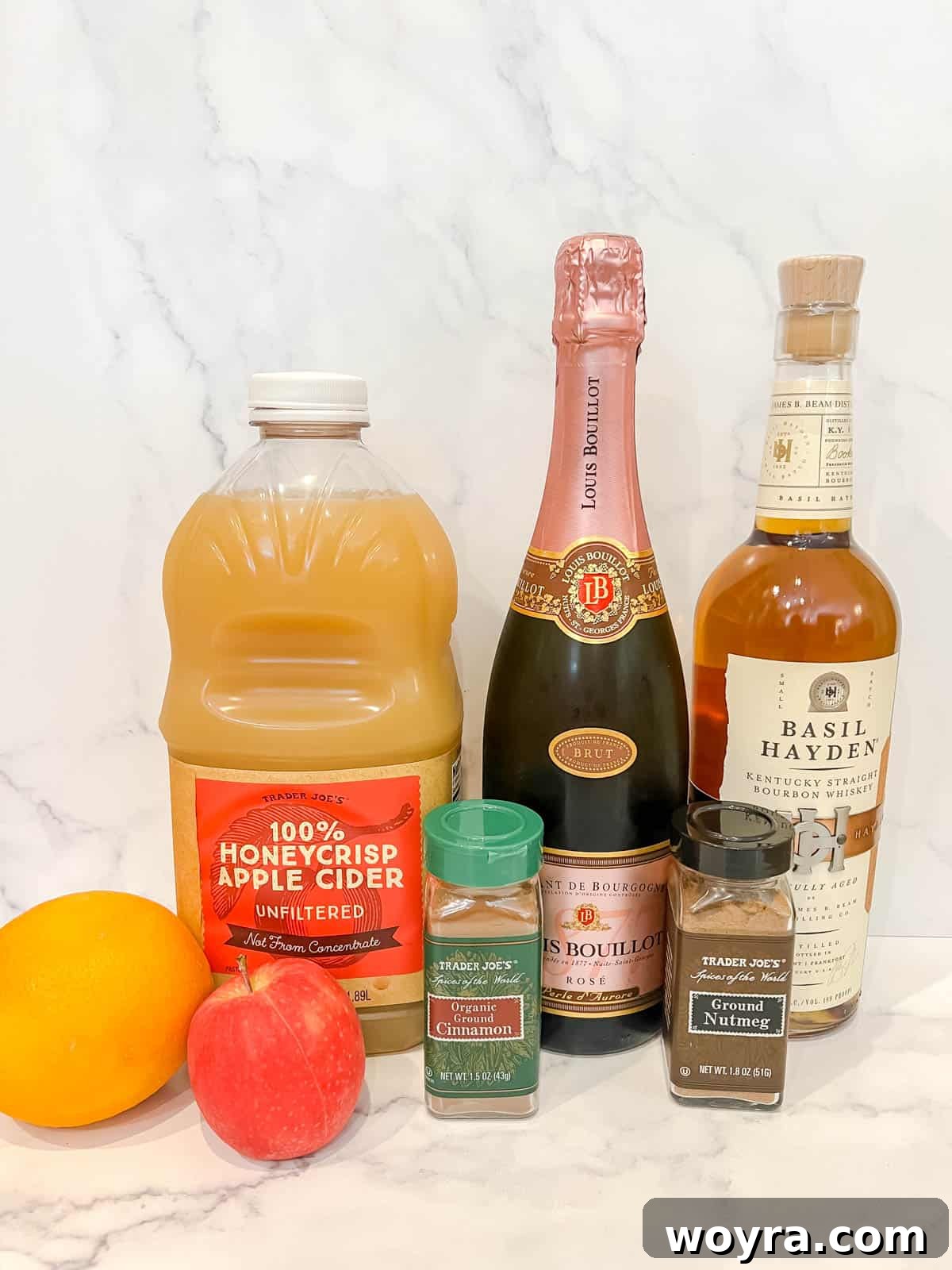 Orchard Ember Elixir 3 A collection of ingredients for an Apple Cider Whiskey Cocktail, including apple cider, Prosecco, whiskey, spices, and fruit slices.