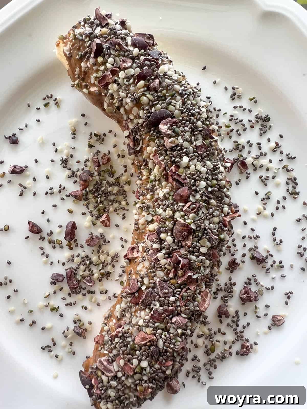 A peeled banana on a plate being coated with almond butter and various seeds.